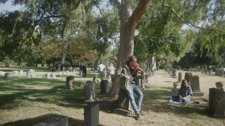 A man is leaning against a tree in a cemetery and is playing the violin and behind him there is a woman taking care of two little boys