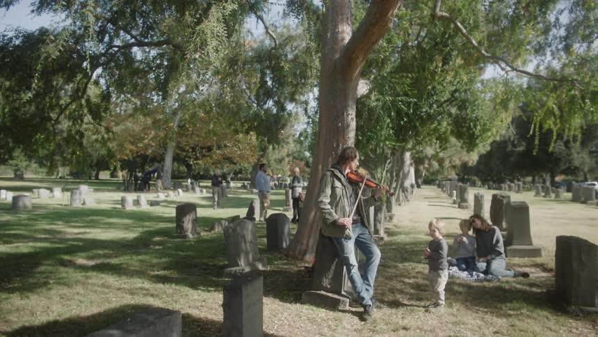 A man is playing the violin while leaning against a tree in a cemetery and there is a child in front of him