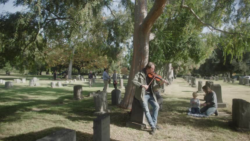A man playing the violin leans against a tree in a cemetery and there is a woman looking after two children behind him