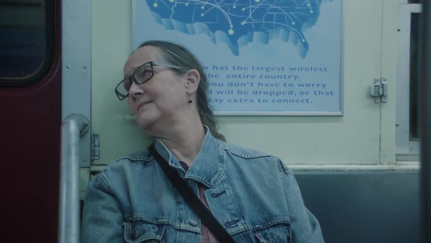 A lady with glasses is sitting on a subway seat and she turns her head to the right and starts laughing