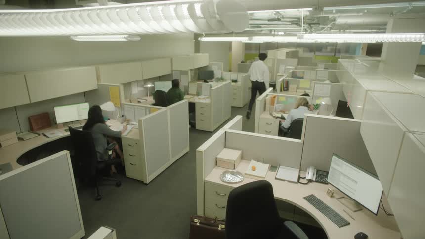 An office with white furniture and three people working and one person is walking in the hallway
