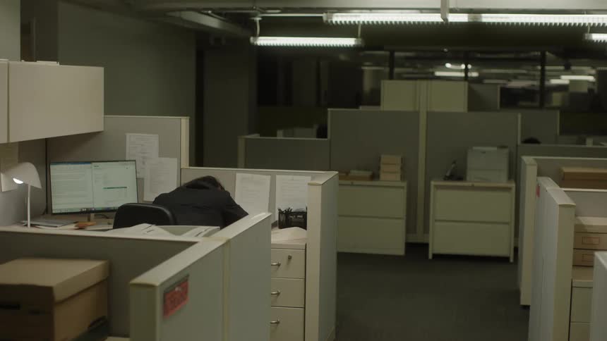 A person wearing a black coat slumped at his desk in a dark office cubicle