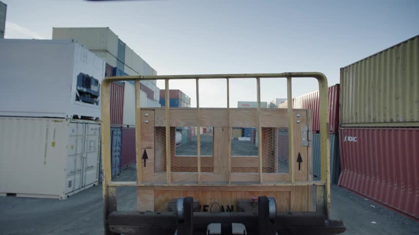 A wooden box being transported by a vehicle with a yellow grille on a path full of containers on its sides