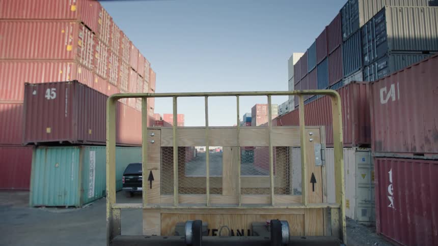 A box being transported by a vehicle with a yellow grille on a path full of containers on its sides