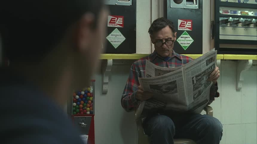 A man sits in a store reading a paper while being watched by another person