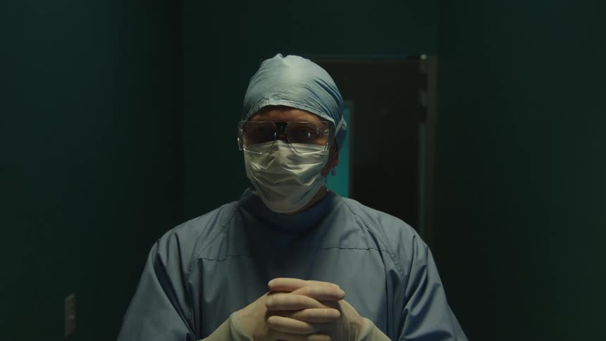 A doctor wearing surgical attire enters a dimly lit hospital room