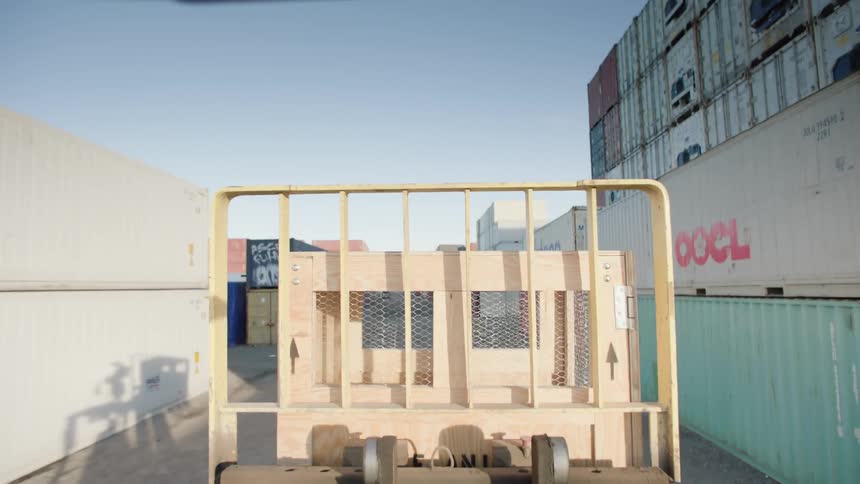 A forklift carrying a wooden box across lanes of shipping containers