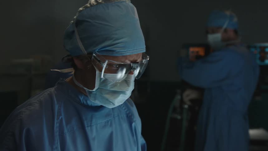 A surgeon performs a medical procedure on a patient while his colleges monitor and discuss it