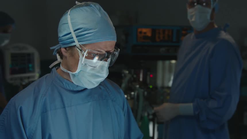 A doctor in a blue coat wearing surgery glasses and a white mask looks straight ahead and shakes his head in a negative sign