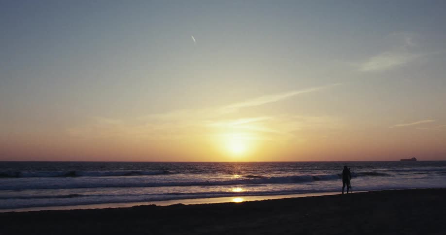 Silhouette of a person with a surfboard walking on the beach sand with the sun on the horizon