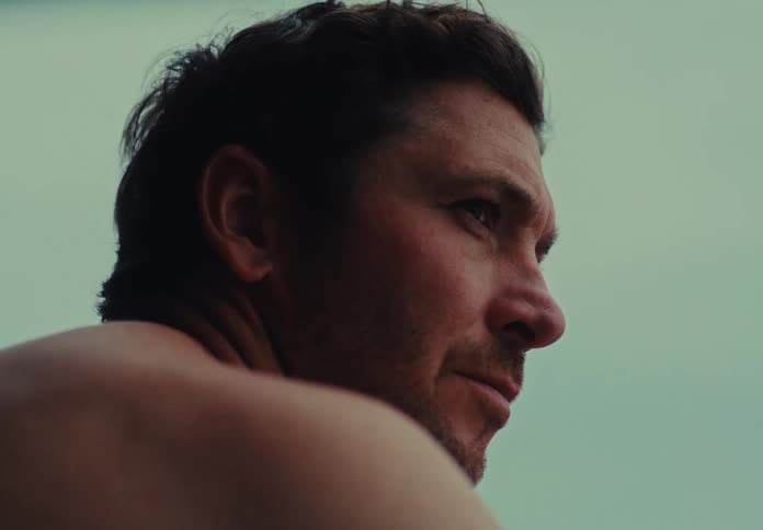A shirtless man looks over his shoulder on the beach