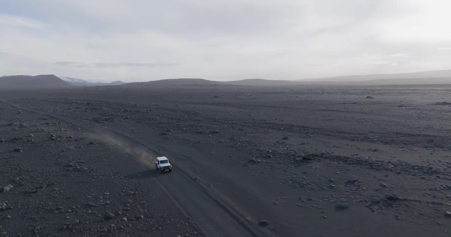 A vehicle travels across a vast barren landscape with mountains in the distance under a cloudy sky