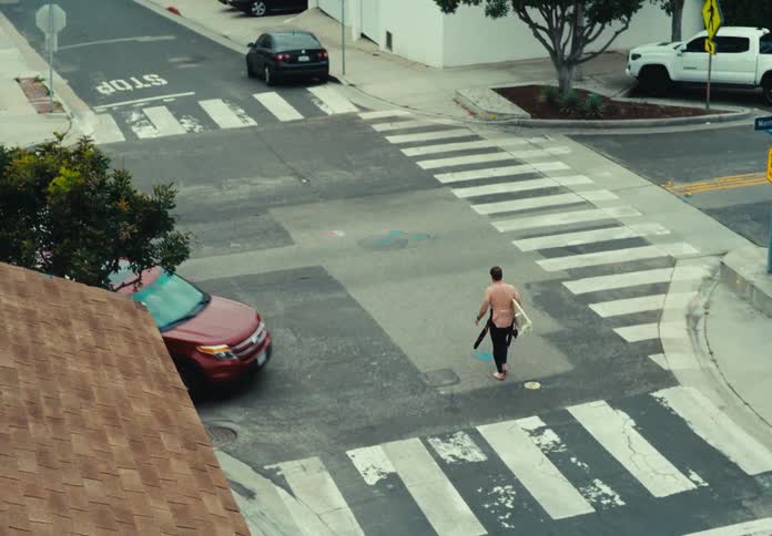 Male surfer carrying a surfboard walking across residential intersection towards the beach
