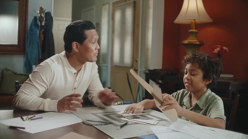 Father and son sitting at the table talking while assembling an airplane out of pieces of cardboard