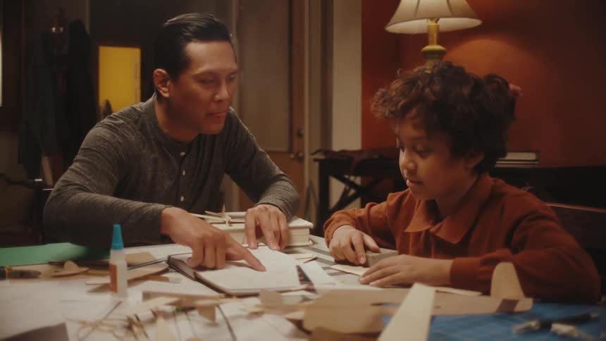 A man and a boy sitting at a table following a blueprint on a notepad to build a wooden toy airplane