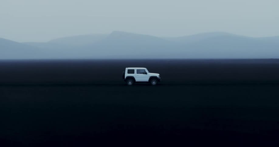 White car traveling on black ground with the silhouette of mountains on the horizon on a foggy day