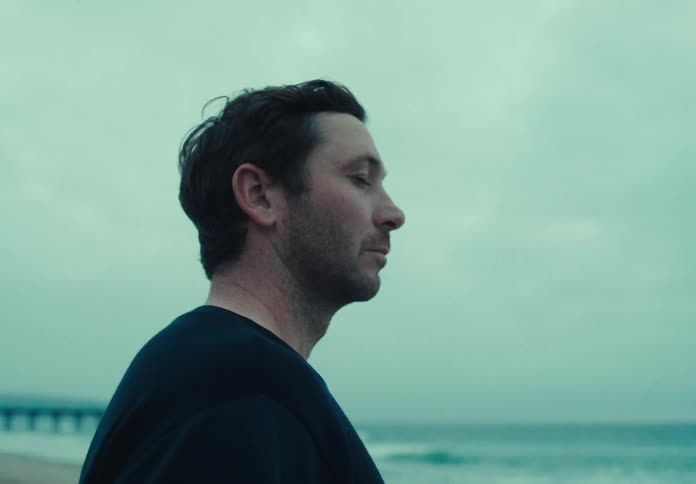 A man in a beach with a long pier looks at the ocean with a pensive look then turns and walks away