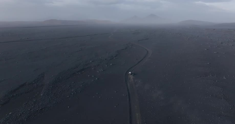 A car on a winding road in a barren dark landscape with distant mountains under an overcast sky