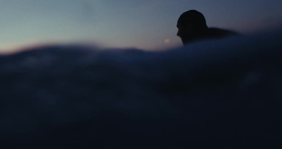 Man with his surfboard swimming in the sea on a purple sky morning