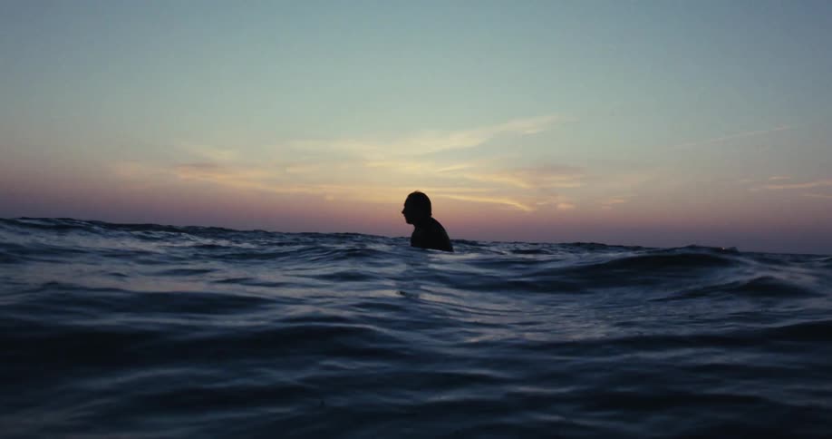 A man in a swimsuit sits on a surfboard in the sea at sunset