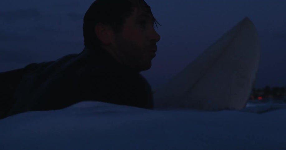 Side silhouette of a mans face with his surfboard in the middle of the sea at night