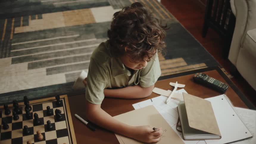 A boy with curly hair writes in a notebook next to a chessboard in the living room