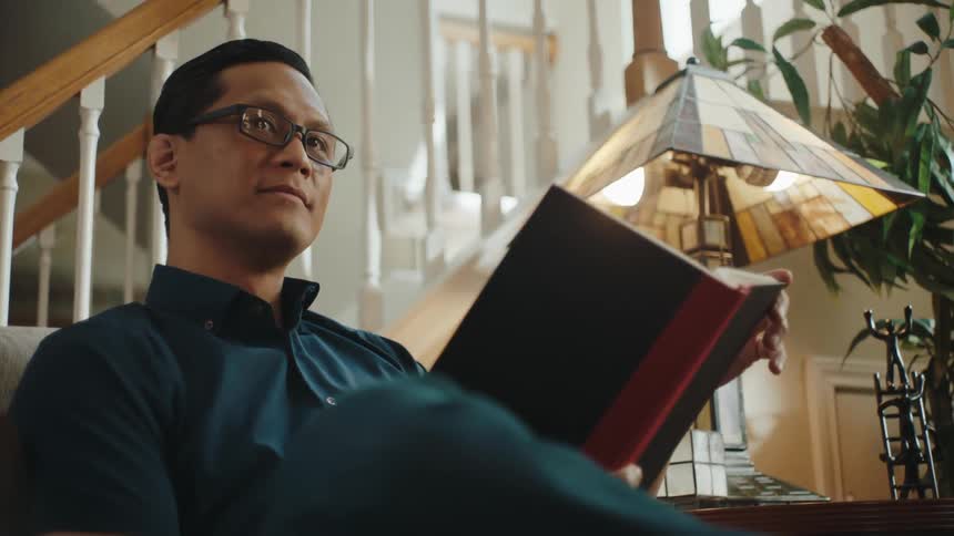 A man wearing glasses a blue shirt and jeans reads a book while sitting in his armchair by a table lamp in front of a staircase