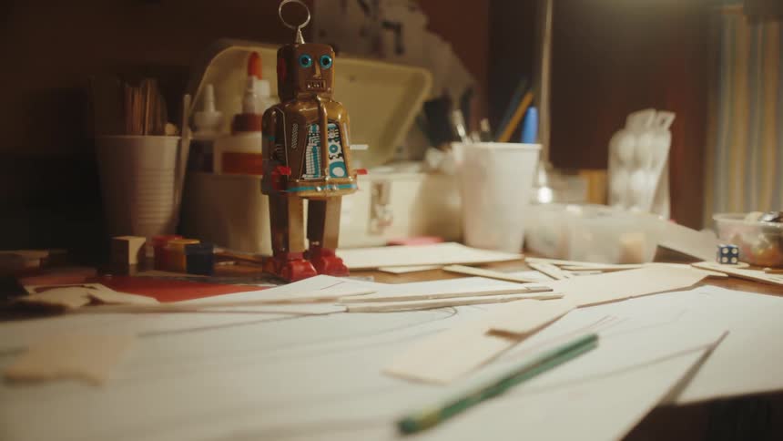 A wooden toy robot over a desk covered in scraps of paper and cardboard