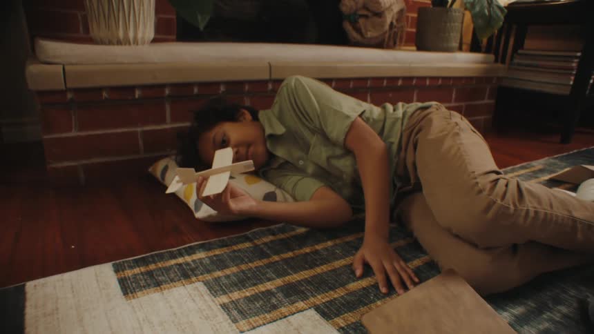 A boy lying on the floor surrounded by scattered papers and books playing with a wooden airplane