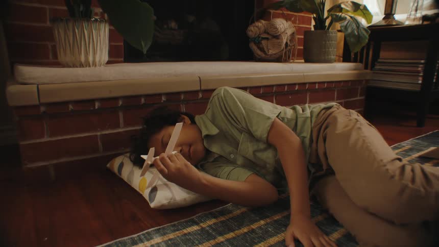 A boy resting in the living room floor plays with a toy paper plane