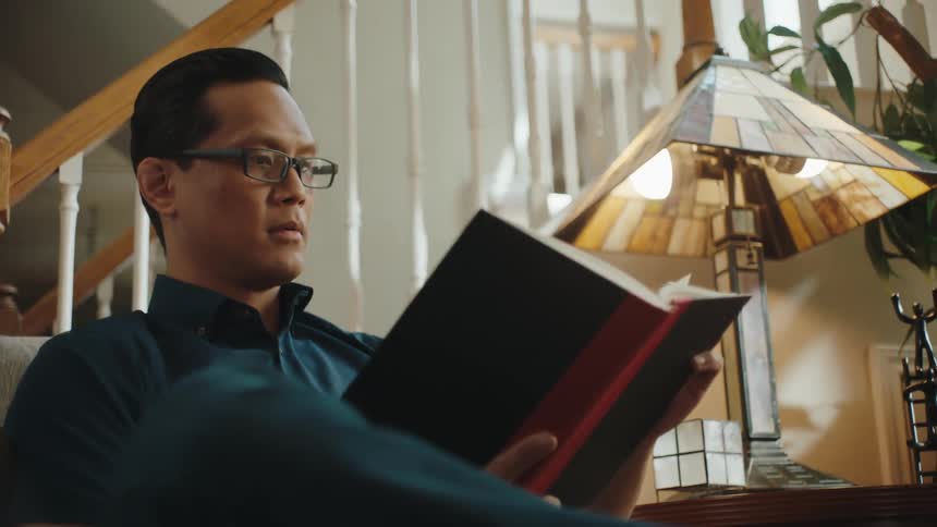 A man sits in a living room reading a book and smiling at someone