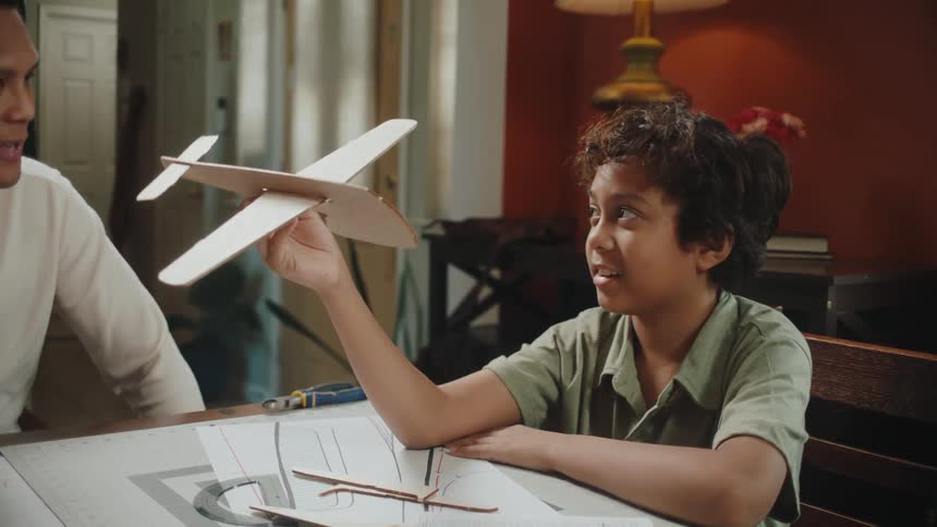 Boy sitting at the table playing with a cardboard plane next to his father
