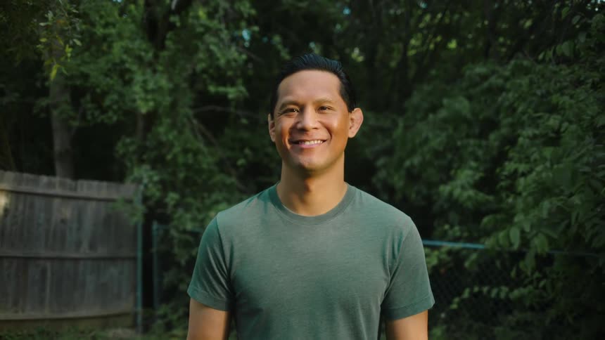 A man smiles and stands in his backyard on a summer day