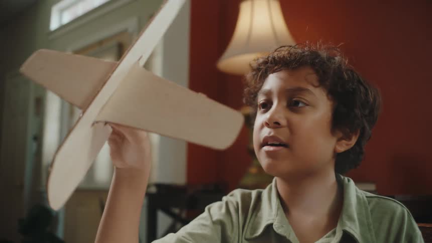 A boy in a green polo shirt sits at a table playing with a wooden airplane