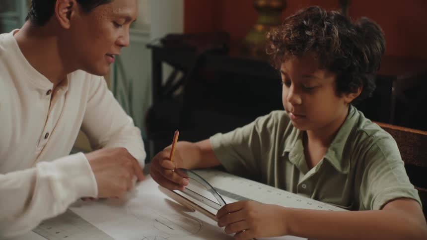 A boy and an adult are sitting at a table and the boy is measuring something with a ruler