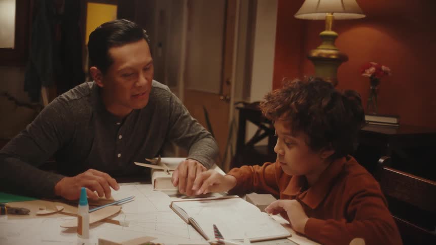 A father and son sitting at a table look at manuals to make a wooden airplane