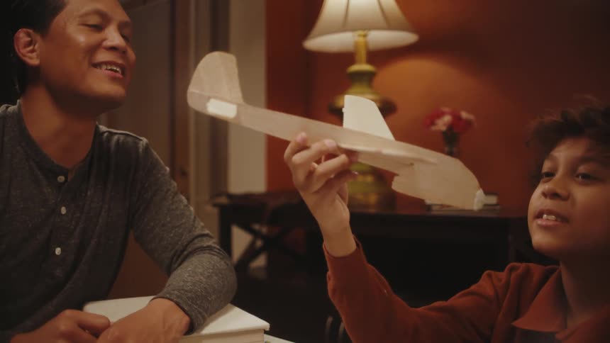 A father and son sitting at a table make a wooden airplane and have a conversation