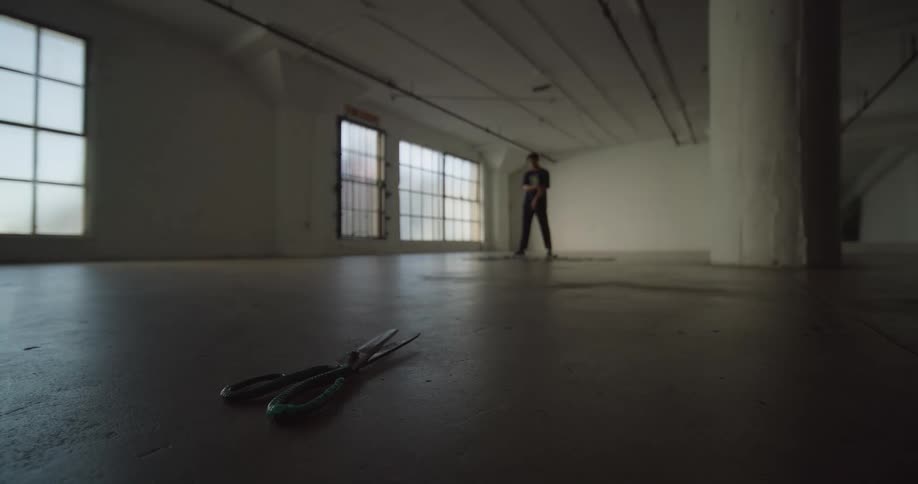 A scissors falling to the floor and there is a person behind who is standing still in an empty room