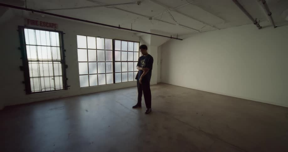 A man has his hand on his elbow and is standing in a totally empty room with large windows