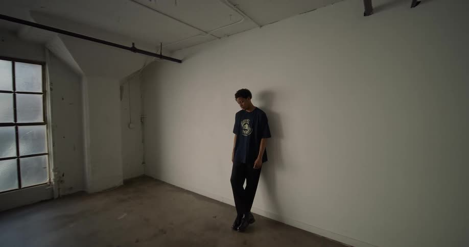 A young man is leaning against a white wall in an empty room he has a thoughtful look on his face