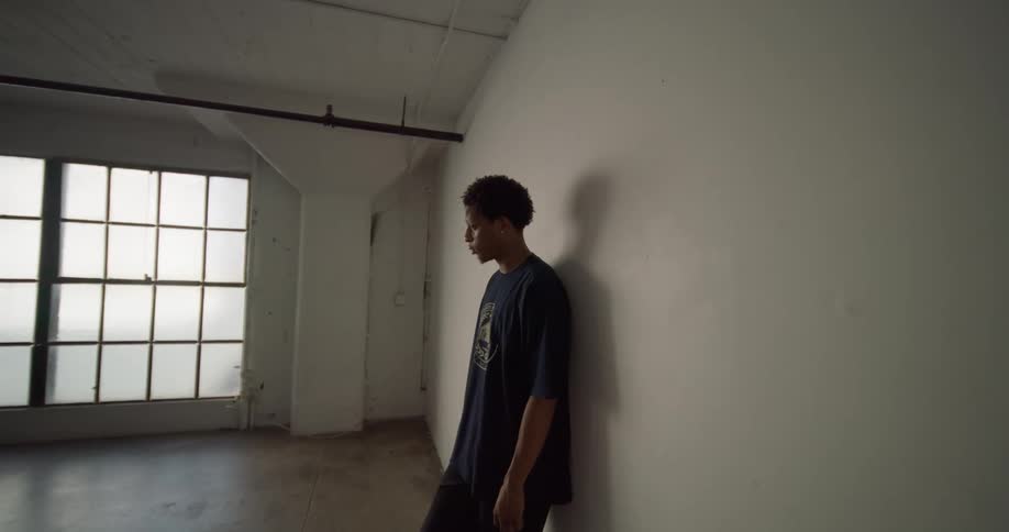 A young man is leaning against a white wall in an empty room with large windows