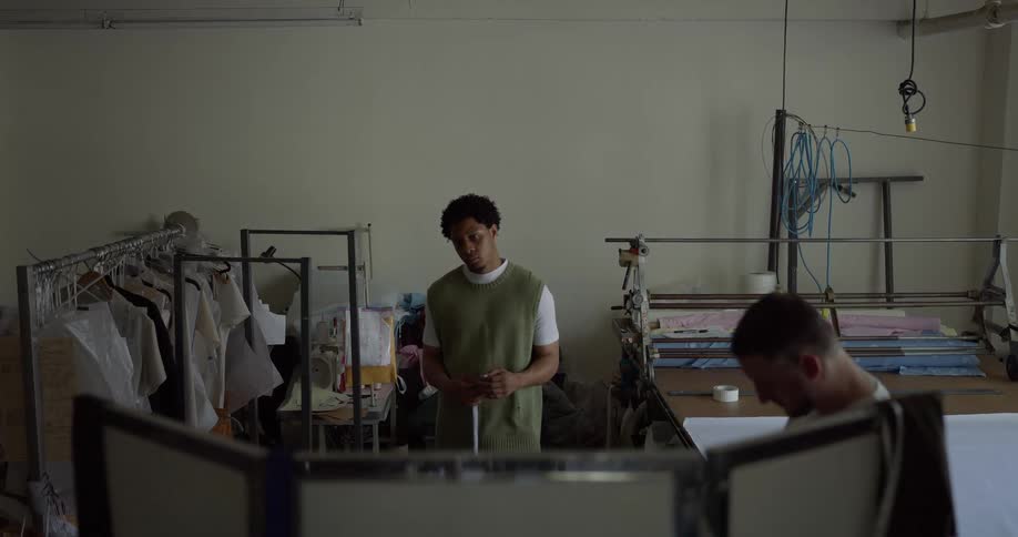 Tailor standing in workshop holds a tape measure while his customer stands near a folding screen