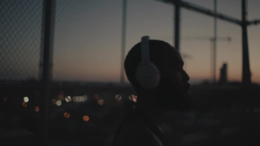 A black man with headphones stands outdoors in a city