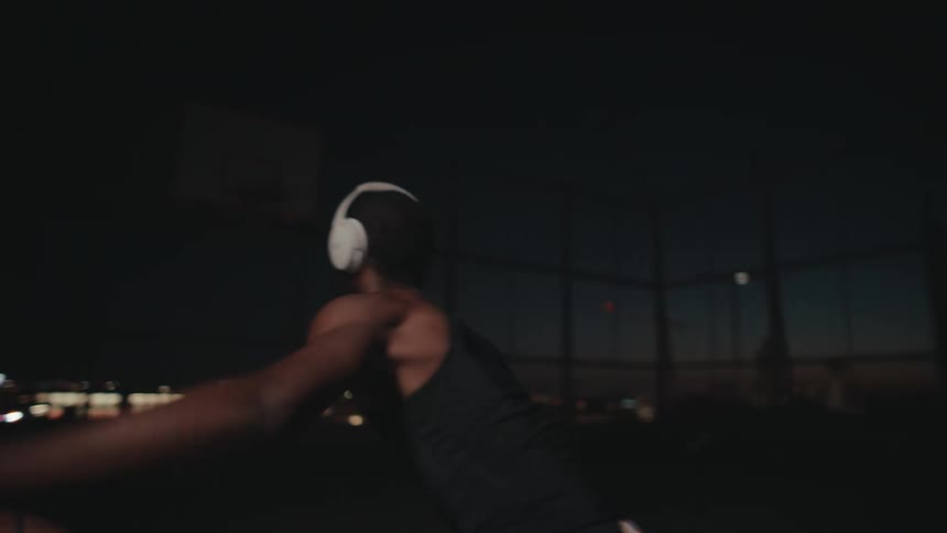 A man wearing white earphones practices basketball at night