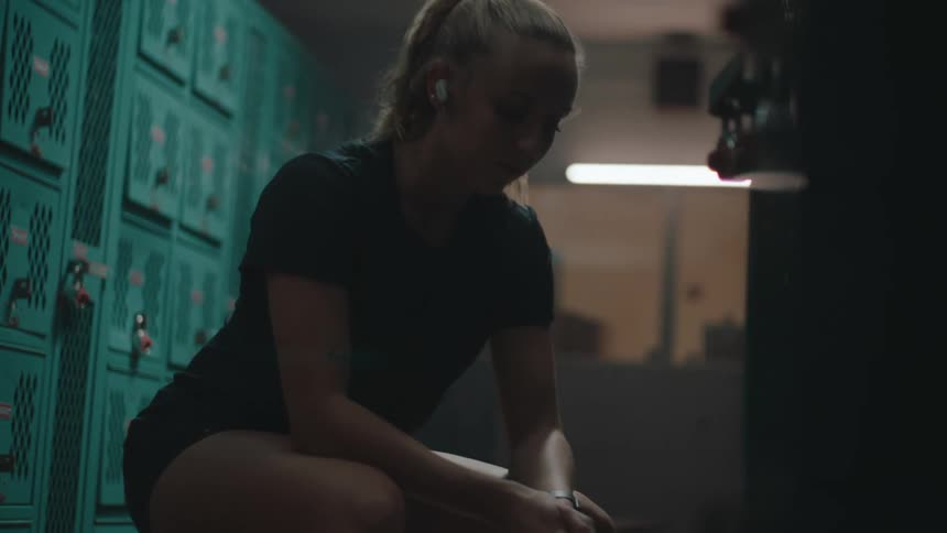 A blonde woman is sitting and moving her leg while wearing headphones and there are lockers around her