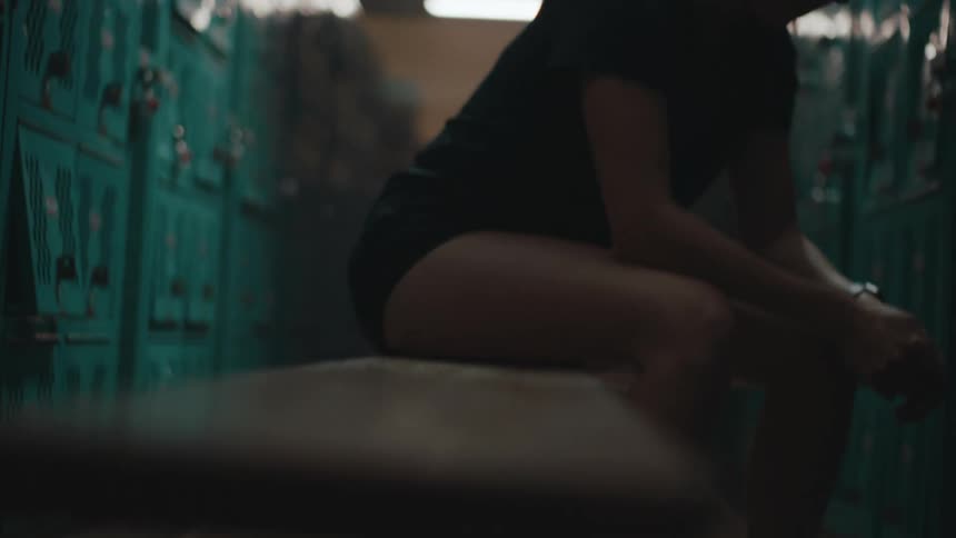 A girl is sitting on a bench and is moving her leg and there are several lockers around her