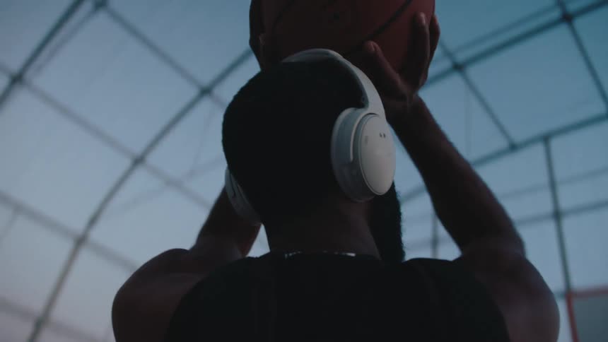 Back view of a man with white headphones shooting a ball into the basketball hoop on a court