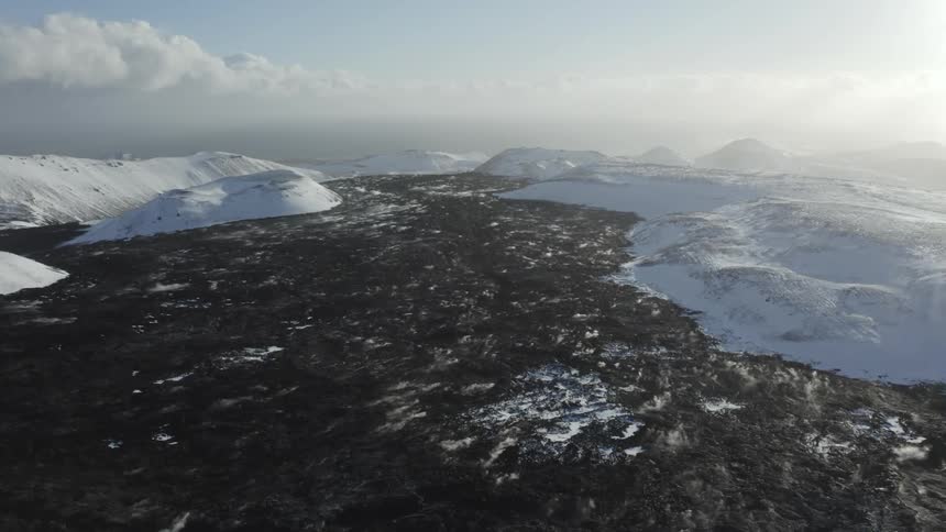 Vapor rises from dark terrain between snow covered mountains on a cloudy day