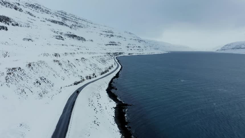 Aerial view of a car driving down a road between a lake and snowy mountain slope