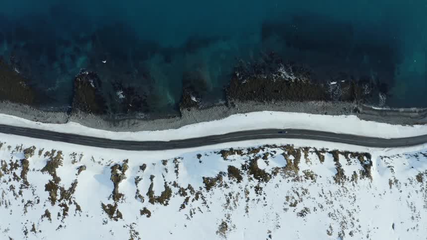 An aerial view of a snowy coastline with waves gently washing over it and a road above it